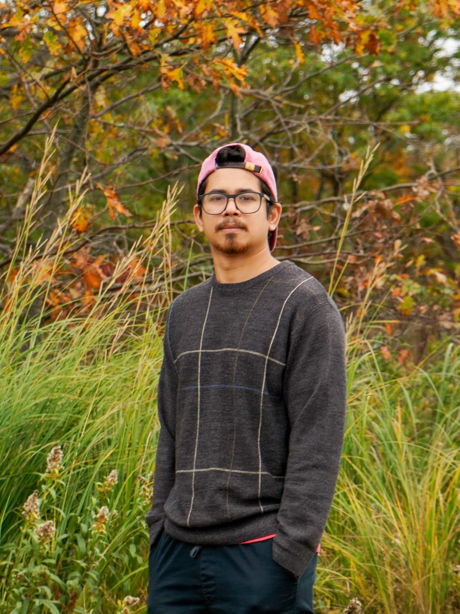 Sundar in glasses, a backwards pink cap, and a grey sweater, standing in autumn grass